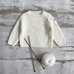 Peut inclure: Cardigan de bébé en tricot crème avec un motif texturé et une fermeture boutonnée. L'image comprend des aiguilles à tricoter et une pelote de laine, suggérant un article fait à la main. Le texte "Little French Knits" est visible.