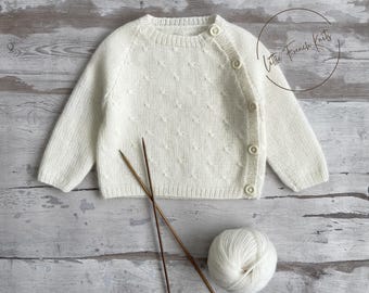 Pode incluir: Cardigã de bebê de malha creme com um padrão texturizado e fecho de botão. A imagem inclui agulhas de tricô e uma bola de lã, sugerindo um item feito à mão. O texto "Little French Knits" é visível.