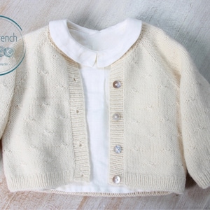 Puede incluir: Un c&aacute;rdigan de punto color crema con un delicado patr&oacute;n calado y botones nacarados. El c&aacute;rdigan se muestra con una camisa blanca de cuello debajo.