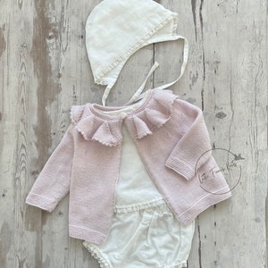 Puede incluir: Un cárdigan de punto rosa para bebé con cuello de encaje blanco, una camiseta de algodón blanca y bombachos con encaje blanco. También se incluye un gorro blanco con encaje. El texto "Little French Knits" es visible en la imagen.
