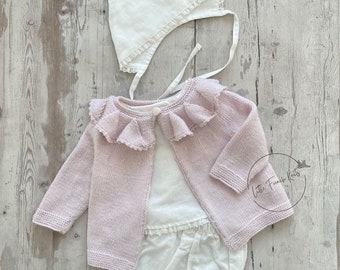 May include: A pink knitted baby cardigan with a white lace trim collar, a white cotton t-shirt, and white lace trimmed bloomers. A white bonnet with lace trim is also included. The text "Little French Knits" is visible in the image.