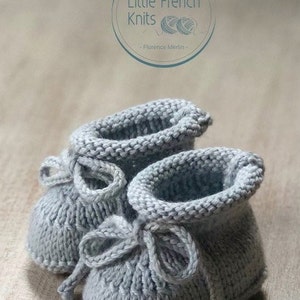 May include: A pair of gray knitted baby booties with a bow on each. The booties are made with a simple stitch pattern and have a soft, cozy look.