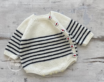May include: A white and navy blue striped knitted baby onesie with red and white buttons. The onesie has a round neckline and long sleeves.