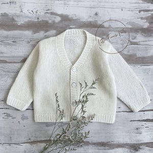 Può includere: Un cardigan in maglia color crema con scollo a V e chiusura con bottoni. Il maglione a maniche lunghe è esposto su una superficie di legno chiaro. Il testo "Little French Knits" è in un cerchio in alto a destra.