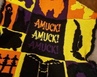May include: A crocheted blanket with a Halloween theme. The blanket is made up of squares with different designs, including a black cat, a ghost, a pumpkin, and a witch's hat. The squares are all in shades of black, orange, yellow, and purple. The blanket has a black border and the words "AMUCK!" are written in white on a purple square.