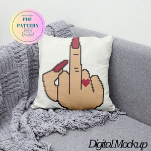 Puede incluir: Una almohada blanca con un diseño de punto de cruz de una mano haciendo el dedo medio con un corazón rojo en el dedo anular. La mano tiene esmalte de uñas rojo. La almohada está en un sofá gris.