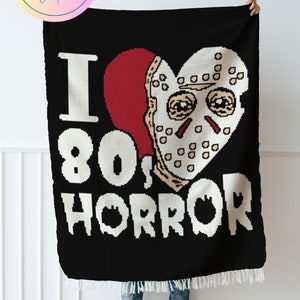 May include: A black and white crochet blanket with a red heart and a pixelated image of a hockey mask. The text "I 80's HORROR" is written in white on the blanket.