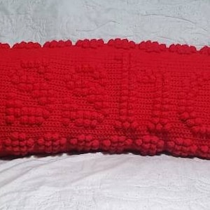 Puede incluir: Una almohada larga de crochet roja con una superficie texturizada y abultada. La almohada tiene un borde festoneado.