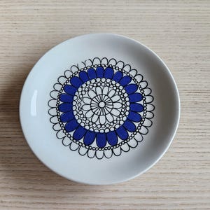 May include: A white ceramic saucer with a blue and black floral design. The design features a circular pattern with blue petals and black outlines.