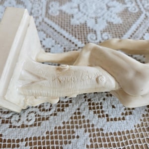 May include: A cream-colored figurine depicting a reclining figure resting against a textured tree trunk base. The sculpture features a smooth, glossy finish and includes a signature etched into the side of the tree trunk base in cursive script.