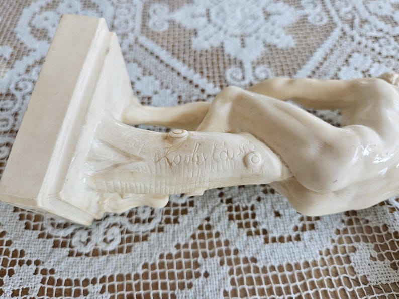 May include: A cream-colored figurine depicting a reclining figure resting against a textured tree trunk base. The sculpture features a smooth, glossy finish and includes a signature etched into the side of the tree trunk base in cursive script.