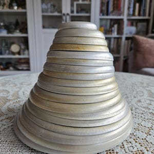 May include: A stack of graduated circular metal pans arranged in a conical shape. The pans are made of a dull, weathered silver-toned metal with visible signs of wear, including faint golden-brown discoloration along the edges of the rims.