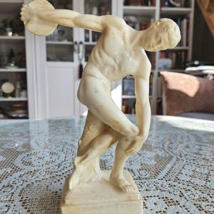 May include: A cream-colored sculpture of a discus thrower in a dynamic athletic pose. The figure is depicted in a forward-leaning stance with one arm extended holding a disc, while the other arm is bent near the knee. It stands on a square base.