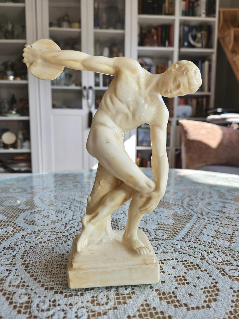 May include: A cream-colored sculpture of a discus thrower in a dynamic athletic pose. The figure is depicted in a forward-leaning stance with one arm extended holding a disc, while the other arm is bent near the knee. It stands on a square base.