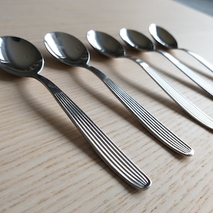 May include: Five silver spoons with a textured handle. The spoons are arranged in a row on a light wood surface.