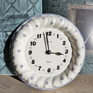 May include: White ceramic clock with blue accents and a white face. The clock has a decorative design around the edge and a quartz movement. The clock is made in Germany.