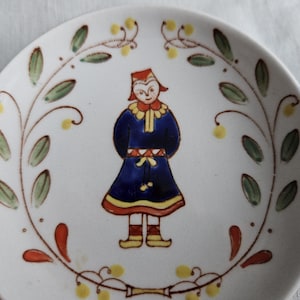 May include: A white ceramic plate with a blue and red folk art design of a person wearing a blue dress and red hat. The plate is decorated with green and yellow floral designs.