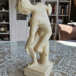 May include: A cream-colored figurine depicting a figure in a forward-leaning pose, holding a circular disc in one hand. The sculpture features a smooth, glossy finish, stands on a rectangular base, and is supported by a tree trunk-like structure.