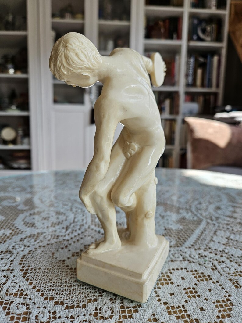 May include: A cream-colored figurine depicting a figure in a forward-leaning pose, holding a circular disc in one hand. The sculpture features a smooth, glossy finish, stands on a rectangular base, and is supported by a tree trunk-like structure.