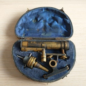 May include: A vintage brass microscope with a blue velvet lined case. The microscope has a long brass tube with a focusing knob and a small brass stand with a screw adjustment.