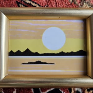 Könnte beinhalten: Ein gerahmtes Kunstwerk, das eine stilisierte Sonnenuntergangsszene mit einer großen weißen Sonne, schwarzen Bergen und einem gelben und orangefarbenen Himmel darstellt.