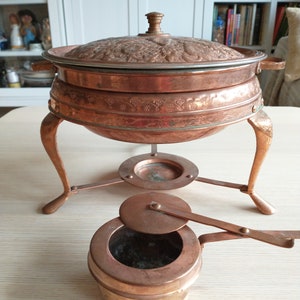 Könnte beinhalten: Eine Kupfer-Chafing Dish mit einem verzierten Design und einem Deckel. Die Schale steht auf einem Ständer mit drei Beinen und einem kleinen Kupferbrenner.