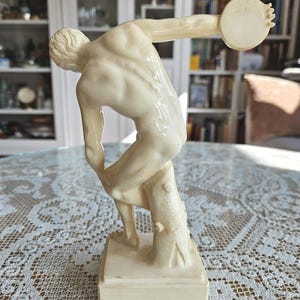 May include: A cream-colored figurine depicting a male athlete in a dynamic pose, preparing to throw a discus. The figure is captured in a mid-motion stance, leaning forward with one arm extended backward, supported by a tree stump on a square base.