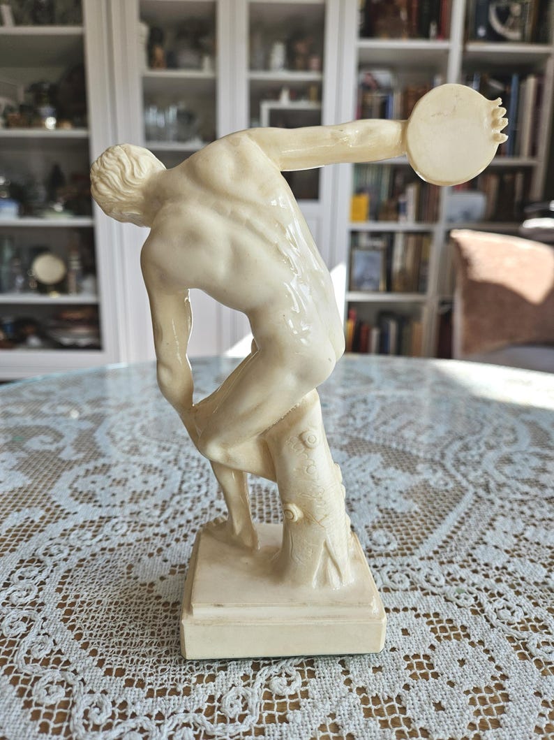 May include: A cream-colored figurine depicting a male athlete in a dynamic pose, preparing to throw a discus. The figure is captured in a mid-motion stance, leaning forward with one arm extended backward, supported by a tree stump on a square base.
