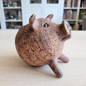 以下が含まれることがあります： テクスチャ加工された表面と黒いアクセントが付いた、茶色のセラミック製のブタの置物。ブタは片方の目がみえており、3本の足で立っています。