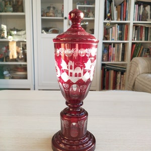 Può includere: Un vaso di vetro rosso con un disegno inciso bianco di un edificio e alberi. Il vaso ha una base rotonda e un coperchio.