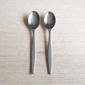 May include: Two silver spoons with rounded bowls and long, slender handles. The spoons are placed side by side on a light wood surface.