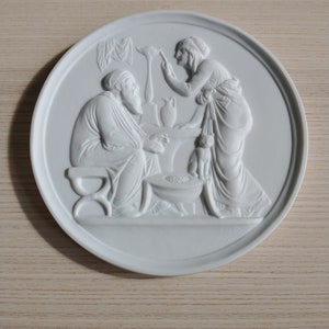 Op de afbeelding: Een wit keramiek bord met een reliëfdesign dat een man en een vrouw in een scène uit de Bijbel voorstelt. De man zit en de vrouw staat, beiden dragen gewaden. De scène speelt zich af in een kamer met een tafel en een vaas.