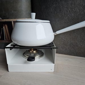 May include: White enamel fondue pot with a long handle, sitting on a white metal stand with a burner. The pot has a lid and is designed for melting chocolate or cheese.