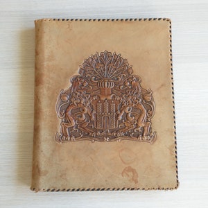 May include: A brown leather journal with a decorative embossed design of a coat of arms featuring a castle and two lions. The journal has a stitched border.