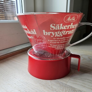 Puede incluir: Un sistema de preparación de café Melitta rojo con un cono de filtro de plástico transparente. El cono tiene texto en sueco y un asa. La base es de color rojo a juego.