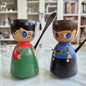 May include: Two wooden egg cups shaped like people with painted faces and clothing. The egg cup on the left is wearing a green and red outfit, while the egg cup on the right is wearing a blue and black outfit.