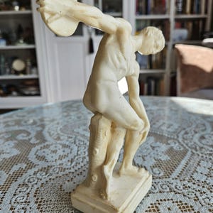 May include: A cream-colored figurine depicting a figure in a dynamic athletic pose, holding a circular disc high above the head. The figure is leaning against a textured tree stump support, set upon a rectangular base, with a smooth finish.