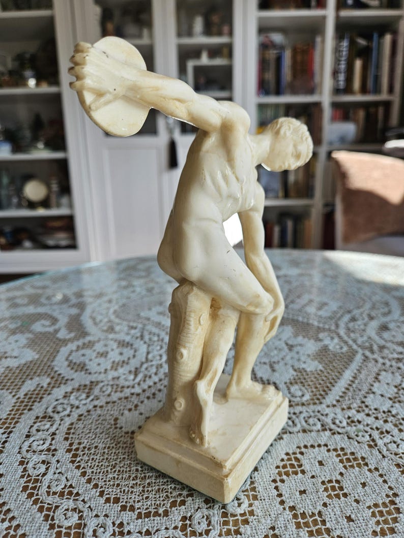 May include: A cream-colored figurine depicting a figure in a dynamic athletic pose, holding a circular disc high above the head. The figure is leaning against a textured tree stump support, set upon a rectangular base, with a smooth finish.
