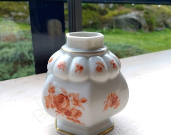May include: A white porcelain vase with a hexagonal base and a floral design. The vase has a scalloped top and is decorated with orange roses and leaves.