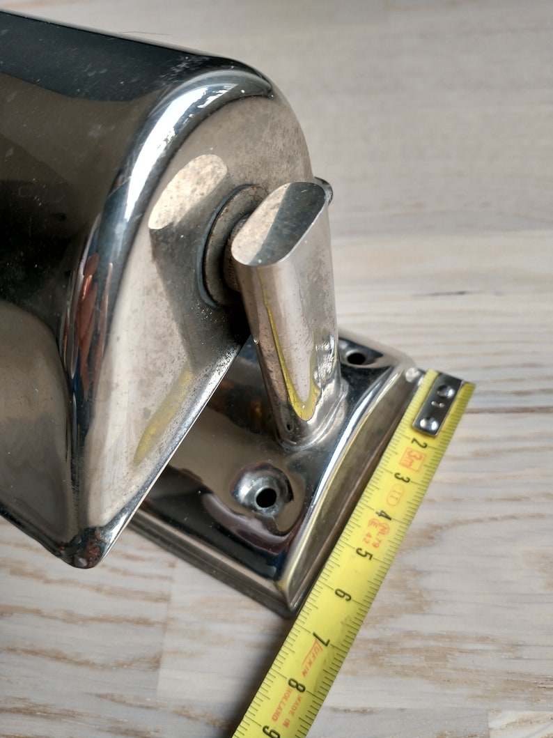 May include: Chrome-plated wall-mounted bathroom tissue holder with a rounded rectangular base and a curved top. The holder is measured with a yellow tape measure.
