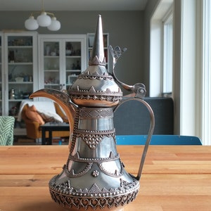 Puede incluir: Una cafetera árabe de plata con intrincados detalles decorativos y una tapa con bisagras. La cafetera tiene un pico y un asa.