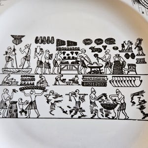May include: White ceramic plate with a black, ancient Egyptian-style illustration. The design depicts figures engaged in various activities, including food preparation and carrying goods. The plate's rim is visible, and the overall aesthetic is historical.