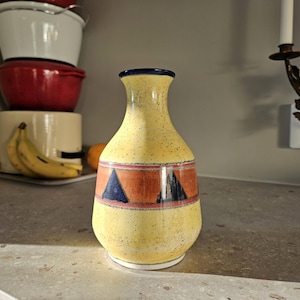 Op de afbeelding: Een gele keramische vaas met een blauw en rood geometrisch ontwerp. De vaas heeft een gespikkeld glazuur en een smalle hals.