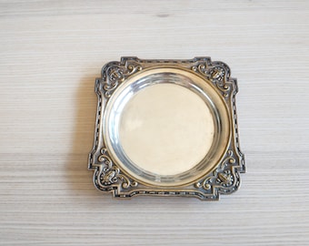 May include: A silver-toned square tray with an ornate design around the edge. The tray has a smooth, reflective surface.
