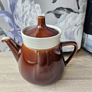 Könnte beinhalten: Eine braune Keramik-Teekanne mit einem weißen Band in der Mitte und einem passenden Deckel. Die Teekanne hat einen gebogenen Ausguss und einen Henkel. Der Hintergrund umfasst ein Blumenmuster und eine Holzoberfläche.