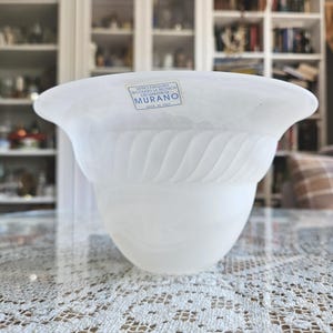 May include: A white, opaque glass bowl with a flared rim and a textured, swirling design around the midsection. The bowl has a label that reads "Murano" and "Made in Italy". The bowl is sitting on a table with a lace tablecloth.