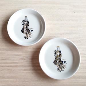 May include: Two white ceramic plates with a gold and blue design of a queen figure. The queen is wearing a crown and a patterned robe.