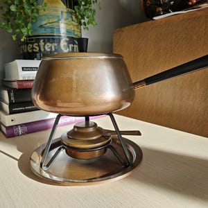 May include: A copper fondue pot with a black handle sits on a black metal stand with a round, copper base. The pot is on a white surface.