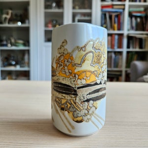 Può includere: Tazza in ceramica bianca con un design astratto marrone, giallo e bianco. Il design presenta una serie di forme e linee sovrapposte.