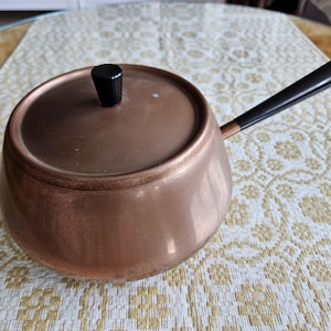 May include: A copper saucepan with a lid and a long black handle. The pot has a rounded shape and a black knob on the lid. The handle is angled upwards. The pot is sitting on a patterned tablecloth.